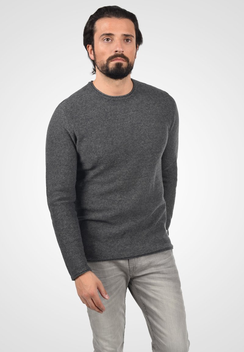 Suéter de cuello redondo gris oscuro con mangas largas, hecho de un tejido texturizado. Combinado con pantalones de mezclilla gris claro. El modelo está de pie contra un fondo neutro.
