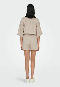 Tenue beige en mélange de lin avec manches larges et taille cintrée, accompagnée d'un short. Portée avec des chaussures blanches, affichant un style décontracté et confortable.