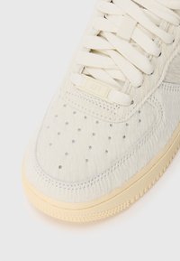 Sleek sneaker branco com parte superior texturizada em pelo curto, perfurações, sola de borracha bege e atacadores bege. Etiqueta de marcação AF1 em destaque na lateral.