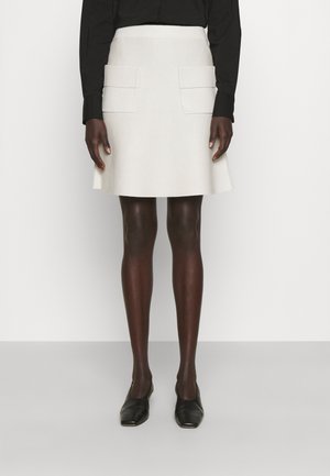 Femme portant une jupe blanche en ligne A avec des poches à rabat à l'avant, un haut noir à manches longues et des chaussures noires à bouts pointus, debout contre un mur blanc.
