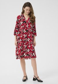 Robe fleurie rouge avec des manches trois-quarts, décolleté en V, au design à volants, avec des motifs de feuilles blanches et noires. Portée avec des chaussures à talons noires.