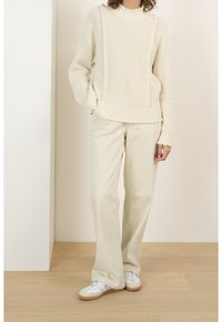Frau in einem cremefarbenen Strickpullover, beigefarbenen, weiten Hosen und weißen Sneakers, die auf einem Holzboden vor einem hellen Hintergrund steht.