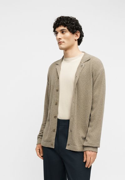 Άνδρας με σγουρά σκούρα μαλλιά που φοράει μπεζ υφασμάτινη ζακέτα με κουμπιά πάνω από κρεμ πουκάμισο και σκούρο παντελόνι, κοιτάζει προς τα πλάγια σε απλό φόντο.