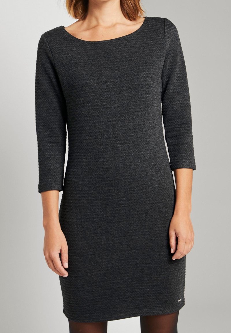 Femme portant une robe cintrée gris foncé texturée avec des manches trois-quarts et un col rond sur un fond clair uni.
