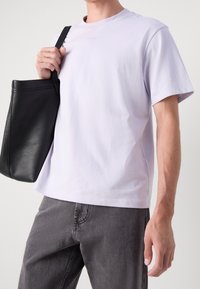 Mężczyzna w gładkiej, białej koszulce z krótkim rękawem i szarych dżinsach, trzymający czarną skórzaną torbę na ramieniu.