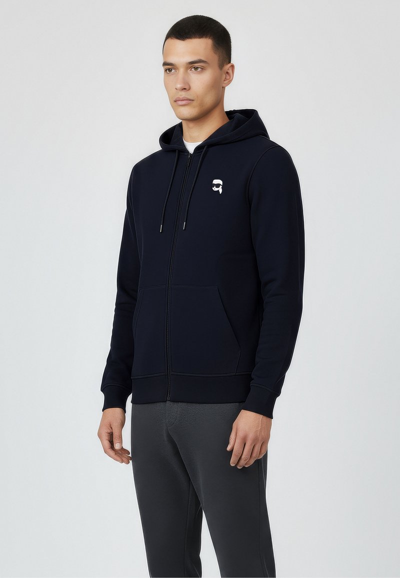 Felpa con zip di colore blu navy realizzata in un tessuto morbido, con tasca frontale e una piccola grafica bianca sul lato sinistro del petto. Include cappuccio con cordini.