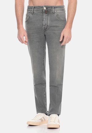 Uomo che indossa jeans grigi a gamba dritta e sneakers bianche, in piedi davanti a uno sfondo bianco.