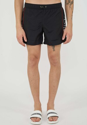 Uomo con pantaloncini da bagno neri con piping rosso e ciabatte bianche Emporio Armani, in piedi su uno sfondo chiaro e semplice.