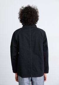 Black jacket with a straight cut, visible stitch detailing, and a collar. Fabric appears sturdy. Back view shows simple, clean lines.