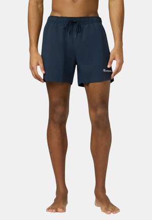 Uomo che indossa pantaloncini da bagno Champion blu navy con coulisse, in piedi a piedi nudi su uno sfondo semplice.