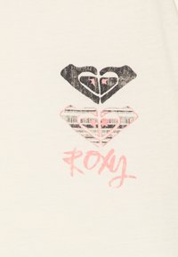 Camiseta Roxy de tejido crema, con tres diseños de corazones en negro y rosa, con un patrón texturizado y un logo escrito a mano.