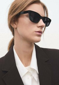 Czarne okulary przeciwsłoneczne typu kocie oko z grubymi oprawkami, gładką teksturą i przyciemnianymi soczewkami. Nosi je z białą koszulą na guziki pod ciemną marynarką.