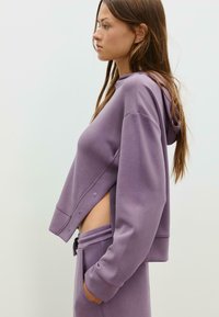 Ljubičasti skraćeni hoodie s vezicom i detaljima na bočnim dugmadi, upareni s odgovarajućim trenirkama. Teksturirani materijal, ležeran dizajn.