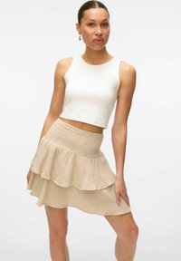 Femme portant un crop top blanc sans manches et une jupe en ruffles beige à plusieurs niveaux, posant avec une main tenant la jupe.