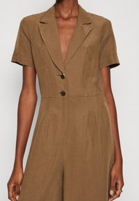 Brun linne jumpsuit med en figursydd överdel, korta ärmar, krage med snitt och två knappar framtill. Slät textur och plisserad midja.