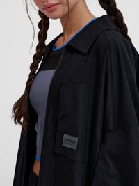 Vrouw met gevlochten haar die een zwart jasje draagt over een aansluitende blauw-zwarte top met een blauwe halslijn, licht naar links gericht.