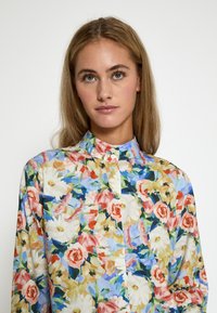 Blommig blus med en flerfärgad design av rosor, prästkragar och gröna blad. Knäppt krage, lång ärm och mjuk tygtextur.