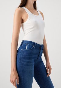 Top sin mangas blanco con escote redondo, combinado con unos jeans de cintura alta en azul oscuro que presentan una textura de denim suave y bolsillos delanteros.