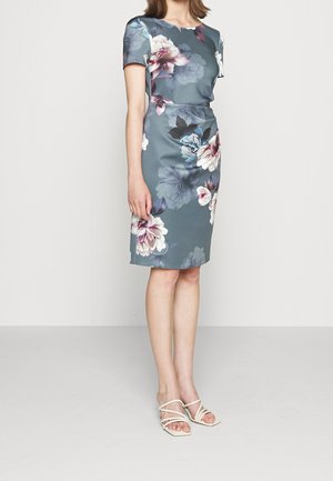 Žena v modrých šatech po kolena s velkými bílými a fialovými květinovými vzory a bílými sandály na podpatku s řemínky, stojící na jednobarevném pozadí.