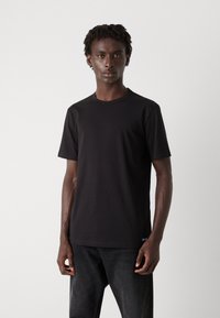 Tricou negru cu maneci scurte, realizat dintr-un material neted, cu decolteu la baza gâtului și un branding subtil în partea stângă jos.