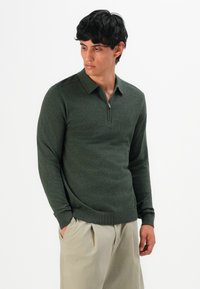 Vyras, vilkintis tamsiai žalią ilgomis rankovėmis užtrauktuku polo megztinį ir šviesiai smėlines kelnes, stovi su rankomis kišenėse, žiūri į kairę.