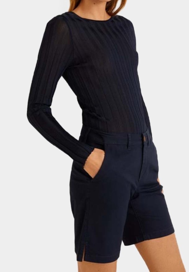 Femme portant un haut côtelé noir à manches longues rentré dans un short tailleur bleu marine foncé taille haute avec des fentes sur les côtés et les mains dans les poches.