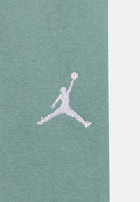 Vit broderad silhuett av en basketspelare som hoppar med en boll på en grön tygbakgrund.