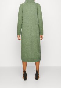 Langes, figurbetontes grünes Strickkleid mit einem hohen Kragen und gerippten Bündchen. Es hat eine glatte Textur und reicht bis zur Mitte der Waden. Schwarze Absätze runden das Outfit ab.
