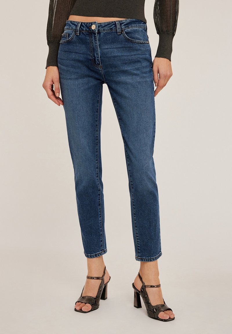 Femme portant un jean bleu droit taille mi-haute et des sandales noires à talons hauts, debout devant un fond uni.