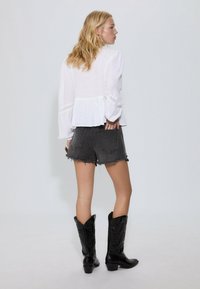 Weiße Bluse mit langen Ärmeln und Spitzenakzenten, kombiniert mit ausgefransten schwarzen Denim-Shorts. Das Model trägt schwarze Cowboy-Stiefel und zeigt eine Rückansicht.