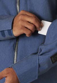 Giubbotto di jeans blu con una texture morbida. Presenta una zip e una tasca per riporre carte sulla manica, accentuata da un'etichetta nera del marchio.