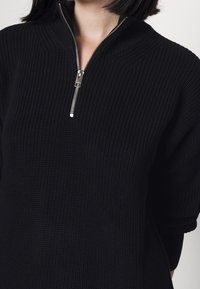 Femme portant un pull noir côtelé en tricot avec un col à demi-fermeture éclair argentée, montrant le cou et le haut du torse.