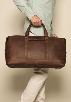 Borsa da viaggio in pelle marrone con due manici robusti, una tasca con zip frontale e dettagli in ottone. Texture liscia, forma rettangolare.
