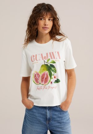Vrouw draagt een wit T-shirt met een kleurrijke illustratie van guavevruchten en bloemen en de tekst "GUAJAVA proef de tropen", gecombineerd met een blauwe spijkerbroek.