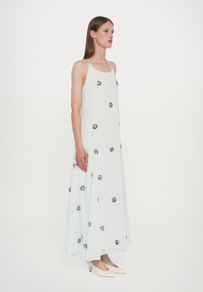 Mujer con un vestido largo blanco sin mangas, con tirantes finos y adornos florales plateados dispersos, combinado con unos zapatos de tacón bajo blancos.