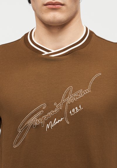 Κοντινό πλάνο ενός ατόμου που φοράει καφέ πουκάμισο με γιακά με λευκές ρίγες και λευκή κεντημένη επιγραφή «Emporio Armani Milano 1.981.»