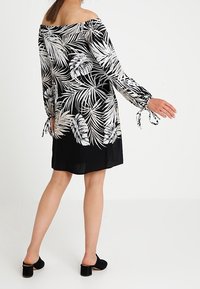 Nainen, jolla on päällä musta olkaimeton mekko, jossa on valkoinen trooppinen lehtikuvio ja solmittavat hihnat, yhdistettynä mustiin korkokenkiin.