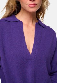 Frau in einem lila Strickpullover mit tiefem V-Ausschnitt und Kragen, der den oberen Brust- und Halsbereich zeigt.