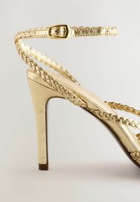 Guld högklackad sandal med flätade remmar, en smal ankle buckle och en slät metallisk yta med en avsmalnande häldesign.