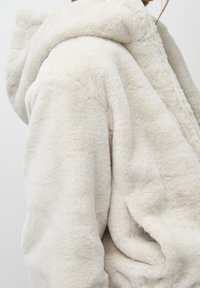 Sweat à capuche couleur crème avec une texture douce et pelucheuse, doté d'une grande capuche, d'une fermeture éclair et d'une coupe décontractée. Tissu lisse et moelleux.