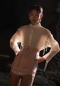 Camicia bianca trasparente con maniche annodate abbinata a una gonna rosa aderente con più tasche e una texture liscia.