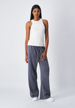 Un débardeur blanc sans manches associé à un pantalon de survêtement ample, gris foncé, avec cordon de serrage. Le pantalon présente une texture douce et un léger effet délavé.