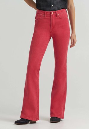 Frau trägt hoch taillierte, ausgestellte rote Jeans in Kombination mit schwarzen, glitzernden Absatzschuhen vor einem einfarbigen, hellen Hintergrund.