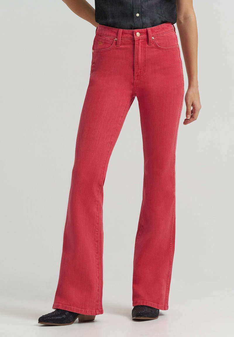 Γυναίκα που φοράει κόκκινο τζιν ψηλόμεσο με καμπάνα, συνδυασμένο με μαύρα γυαλιστερά παπούτσια με τακούνι, σε απλό ανοιχτό φόντο.