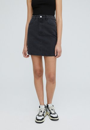 Czarna obcisła spódnica z denimu noszona z czarnym, bezrękawnym topem i czarno-białymi wysokimi trampkami na jasnoszarym tle.