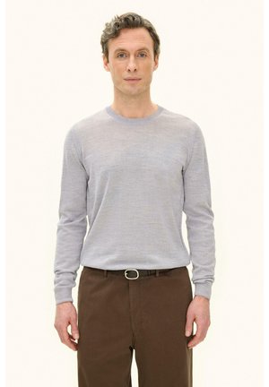 Mężczyzna z krótkimi kręconymi włosami, ubrany w jasnoszary sweter i brązowe spodnie, stoi przodem na jednolitym tle.