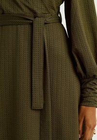 Olivgrön texturerad klänning med en knuten midjebälte och långa puffärmar med rynkade detaljer vid manschetterna.