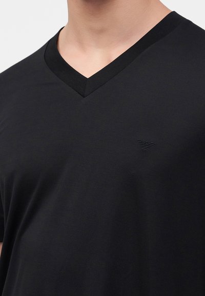 Μαύρο T-shirt με V λαιμόκοψη, φορεμένο από ένα άτομο, με μικρό ανάγλυφο λογότυπο στο αριστερό στήθος.