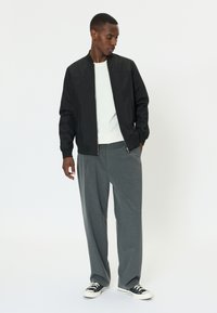 Moški, oblečen v črno bomber jakno, belo srajco, sive široke hlače in črne superge, stoji z rokami v žepih pred belim ozadjem.