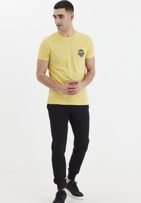 Gul kortærmet t-shirt med rund halsudskæring og et California-motiv. Båret med sorte tætsiddende joggingbukser og sorte sneakers.